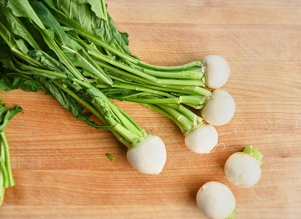 Tokyo turnips on a wooden cutting board, some whole with greens attached and some separated from their greens, leaving only the bulbs.