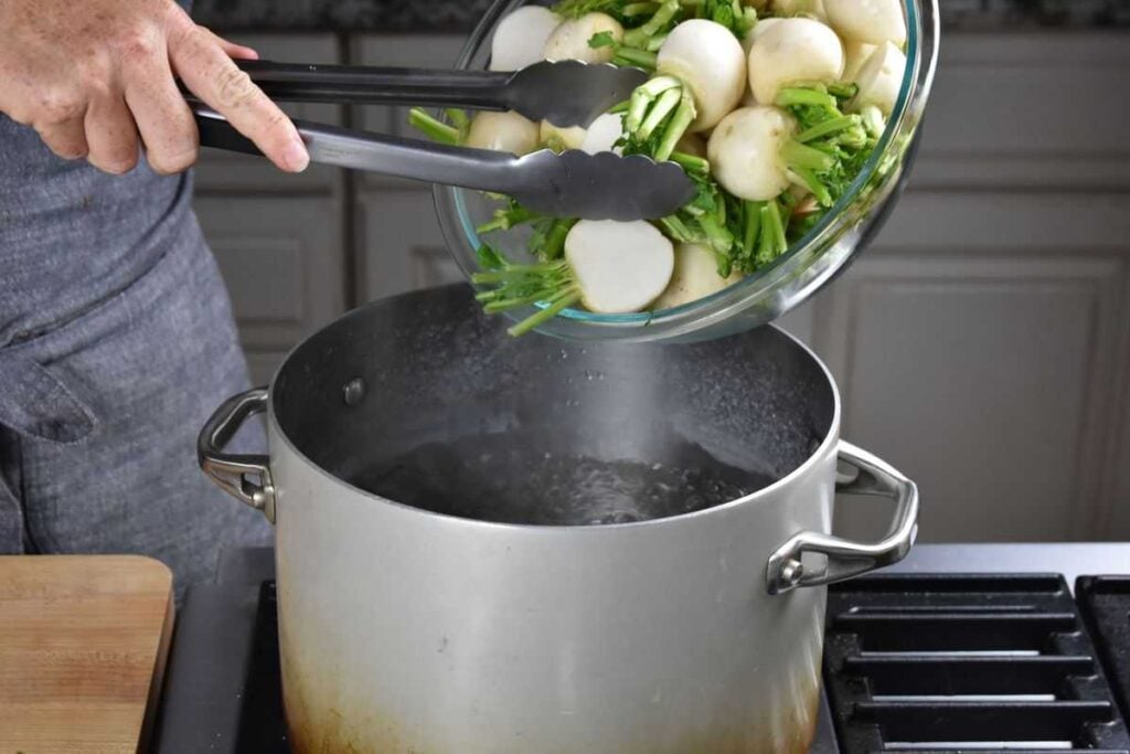 Hand using tongs to place cut Tokyo turnips into a pot of boiling salted water for blanching in a turnip recipe.
