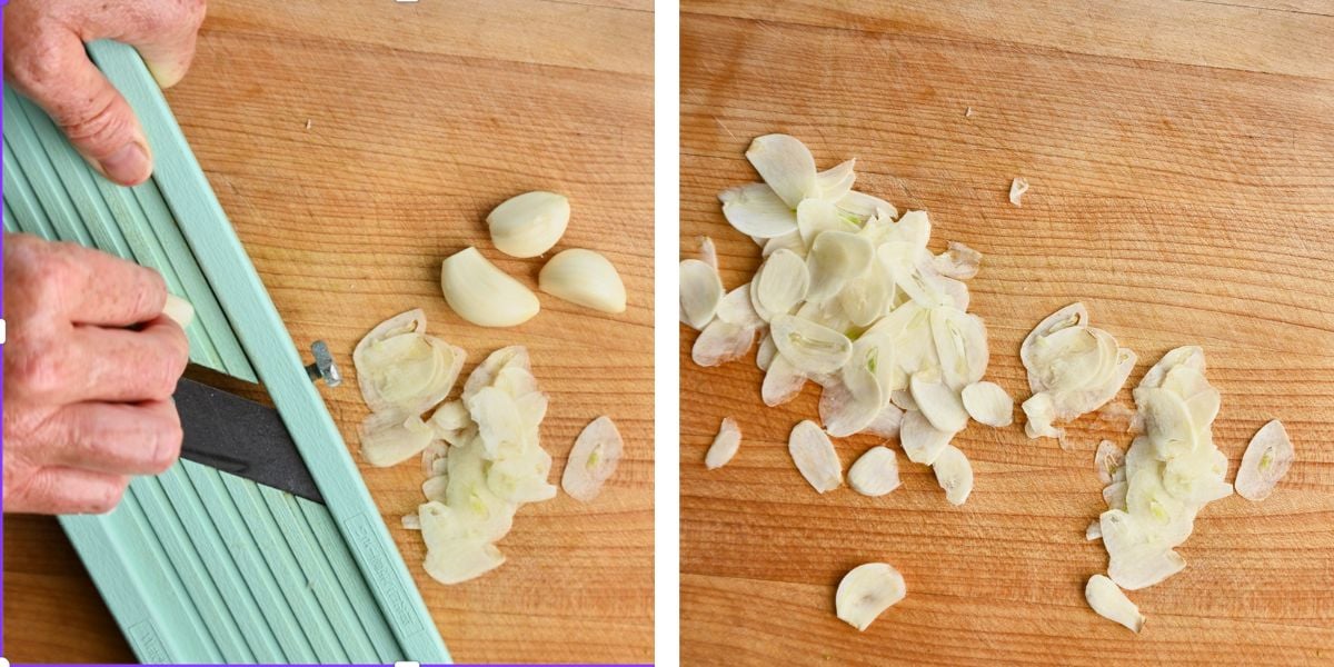 Hand thinly slicing garlic cloves on a Japanese mandolin, with a pile of thinly sliced garlic and whole garlic bulbs arranged on a wooden cutting board.