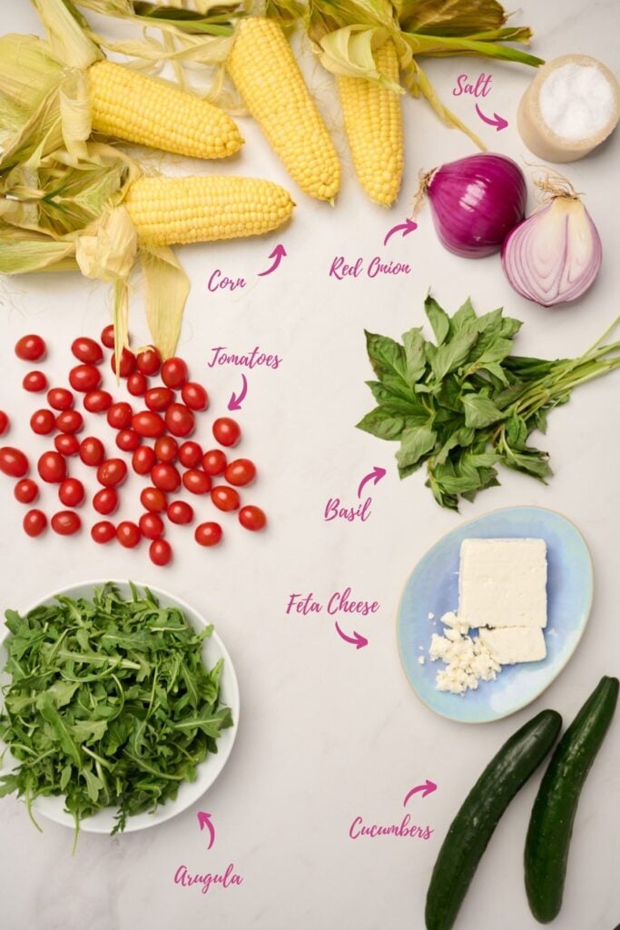 Ingredients for corn salad, including corn, cherry tomatoes, arugula, red onion, basil, cucumbers, feta, and salt.