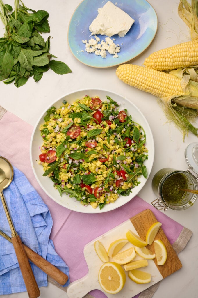 Overhead shot of a fully composed corn salad with purple linen, wooden utensils, corn cob, basil, cut lemons, basil vinaigrette, and feta on the side.