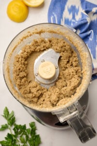 Chunky tahini, garlic, salt, and lemon juice mixture in a food processor, with parsley, lemon, and ikat blue linen in the background.