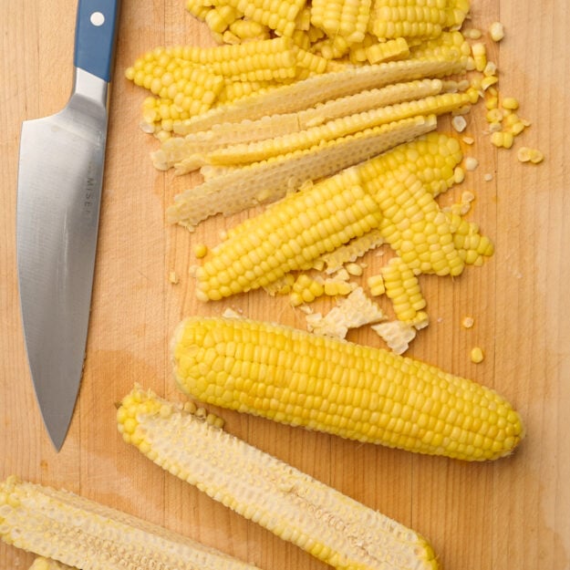 Corn taken off the cob in large pieces on a wooden cutting board with a chef’s knife, surrounded by a pile of corn kernels, bare cobs, and one whole corn cob.