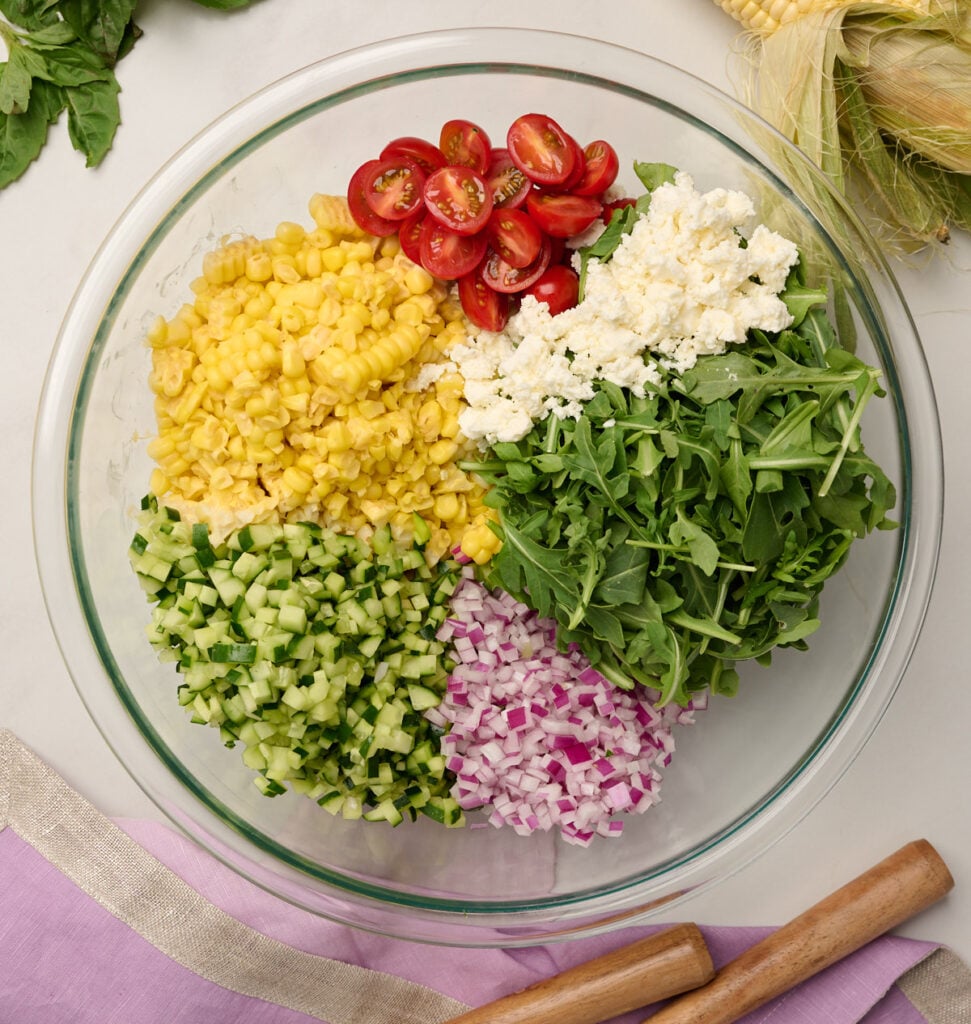 Large glass bowl with corn kernels, sliced tomatoes, arugula, feta, diced red onion, and cucumber, surrounded by a purple linen, wooden utensils, a corn cob, and basil.
