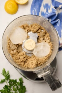Ice cubes being added to a food processor containing tahini mixture, with parsley, lemons, and ikat blue linen in the background.