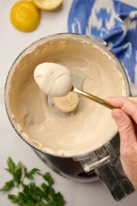 Hand holding a gold spoonful of finished tahini sauce over a food processor, with parsley, lemons, and ikat blue linen in the background.