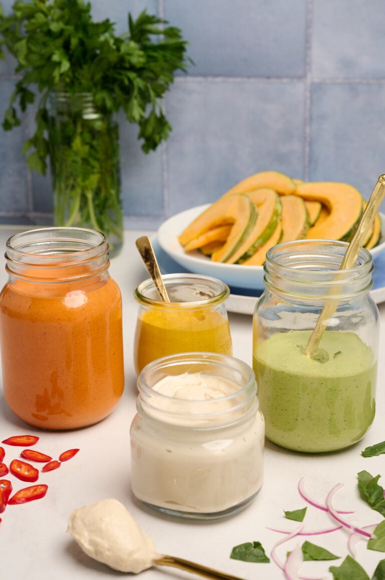 Finished tahini sauce in a jar with a gold spoon, alongside jars of red pepper tahini sauce, turmeric tahini sauce, and herb tahini sauce, with squash, herbs, peppers, and red onion in the background.