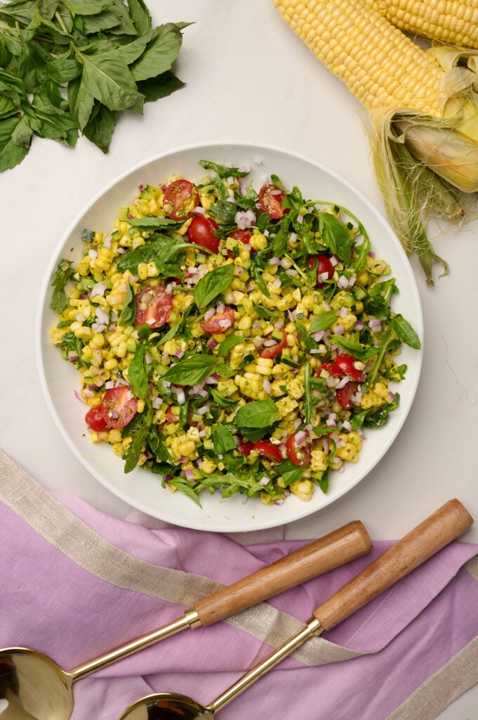 Overhead shot of fully composed and dressed corn salad with purple linen, wooden utensils, a corn cob, and basil.