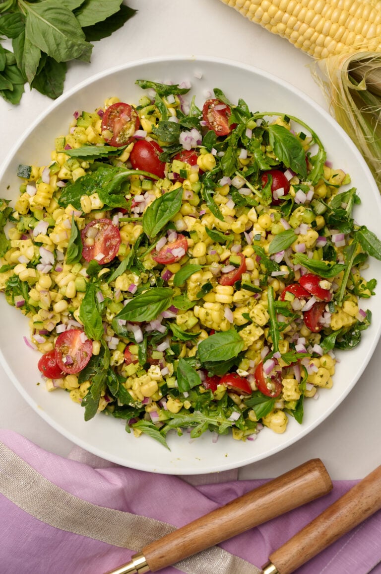 Close-up shot of fully composed corn salad with purple linen, wooden utensils, a corn cob, and basil in the background.