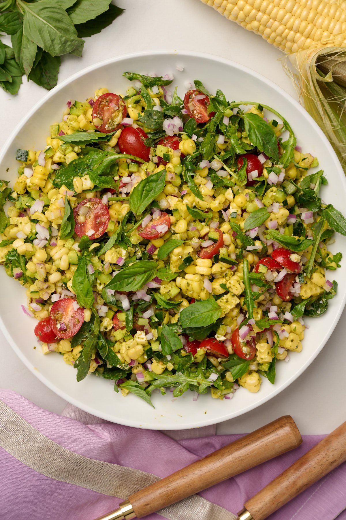Close-up shot of fully composed corn salad with purple linen, wooden utensils, a corn cob, and basil in the background.