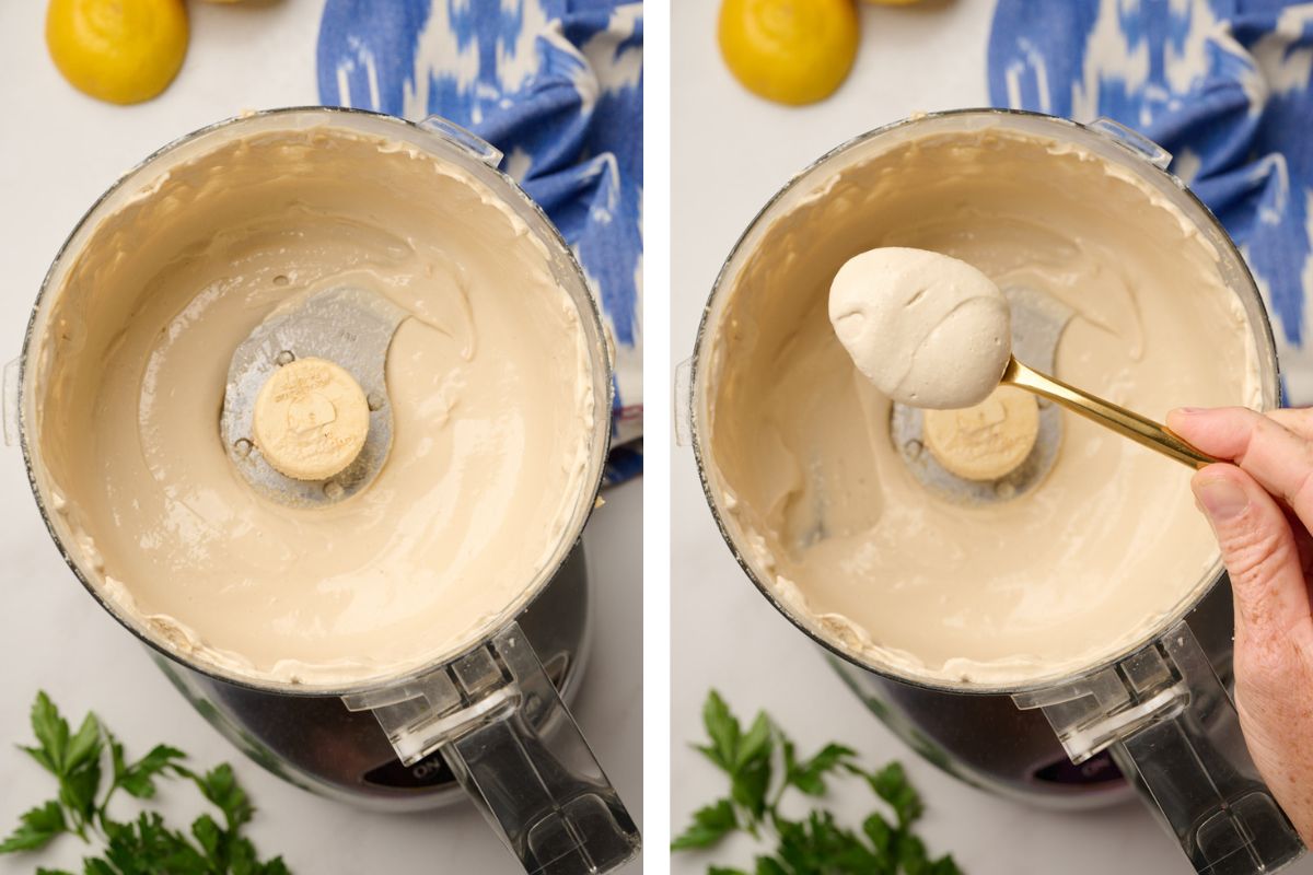 A food processor filled with smooth, creamy tahini sauce, with a hand holding a gold spoonful of the finished sauce, showcasing its perfect consistency. Fresh parsley, lemons, and ikat blue linen are in the background.