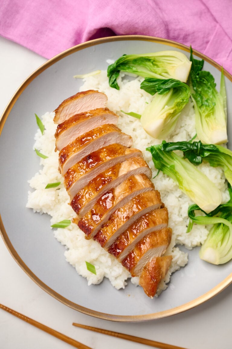 Plated teriyaki chicken breast over steamed rice with baby bok choy and sliced green onions, styled with chopsticks and pink linen.