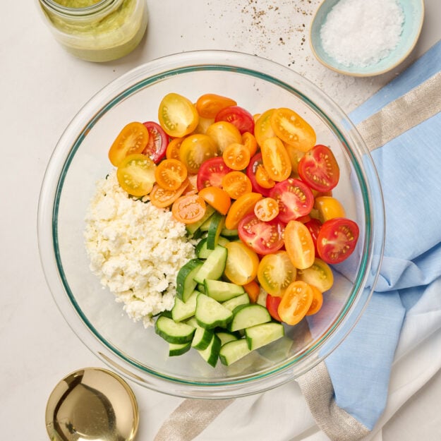 Glass bowl filled with diced cucumbers, tomatoes, and feta cheese, styled with blue linen, avocado dressing, salt, and a gold serving spoon in the background.