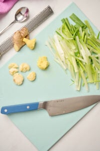 Blue cutting board displaying sliced green onions, smashed garlic cloves, and peeled ginger with a Microplane, silver spoon, and pink linen in the shot.