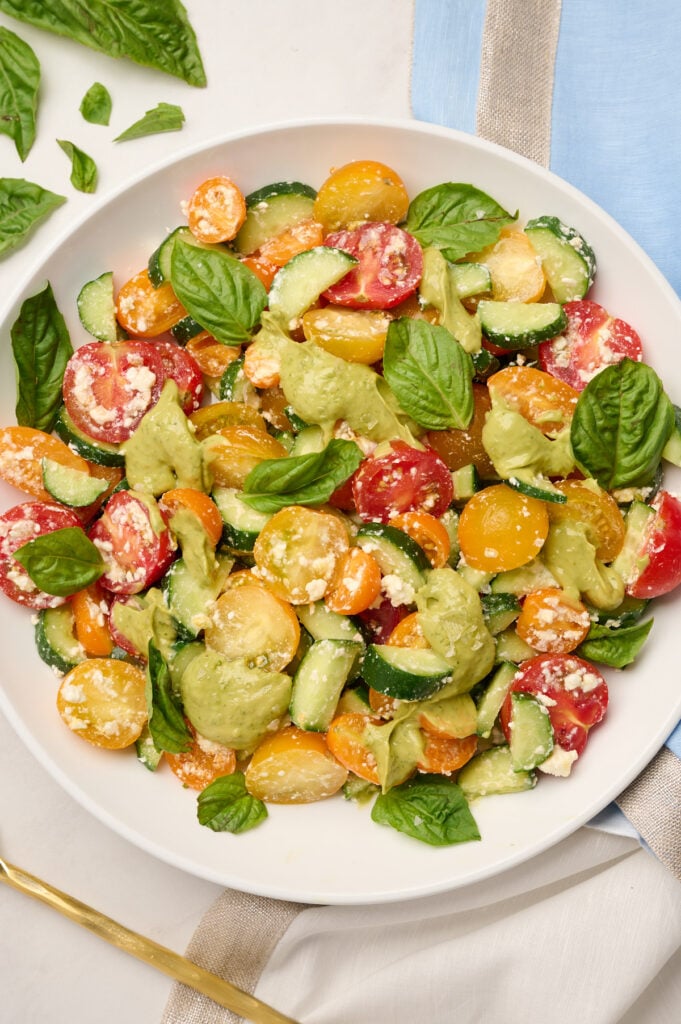 Close-up shot of mixed cucumber tomato salad drizzled with avocado dressing, styled with blue linen, basil, and a gold serving spoon.