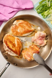 Seared chicken breasts and thighs in a sauté pan, with sliced green onions on a blue cutting board and pink linen in the background.
