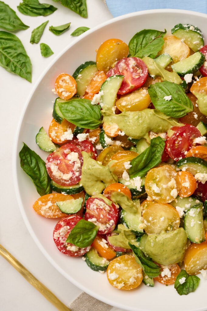 Close-up side shot of fully dressed cucumber tomato salad with avocado dressing, styled with basil, blue linen, and a gold serving spoon.