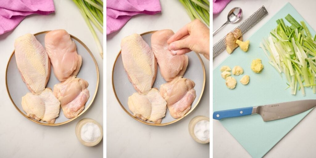 Hand seasoning chicken breasts and thighs with salt and blue cutting board displaying sliced green onions, smashed garlic cloves, and peeled ginger with a Microplane, silver spoon, and pink linen in the shot.