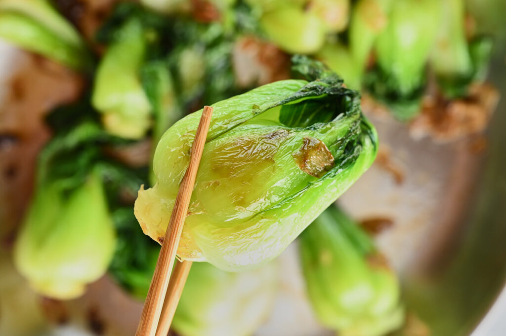 Close-up of chopsticks holding a cooked baby bok choy coated in garlic and soy sauce.