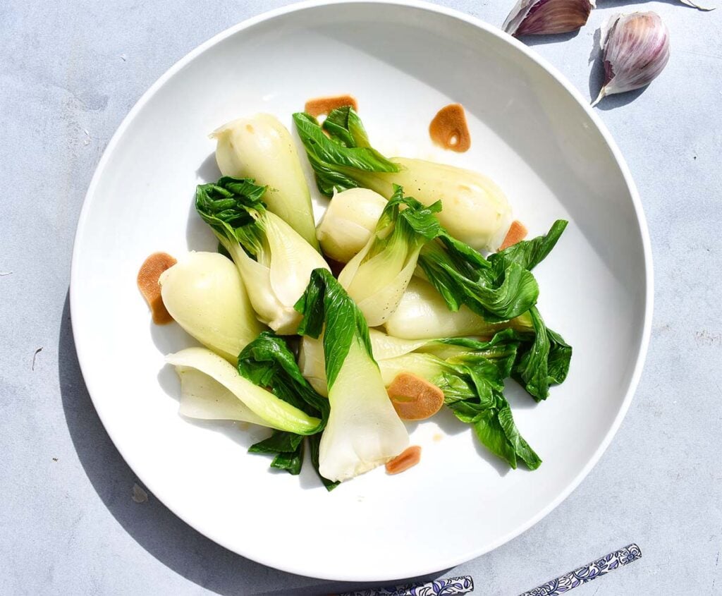 Plated baby bok choy with browned garlic in a white bowl, styled with chopsticks and garlic cloves.