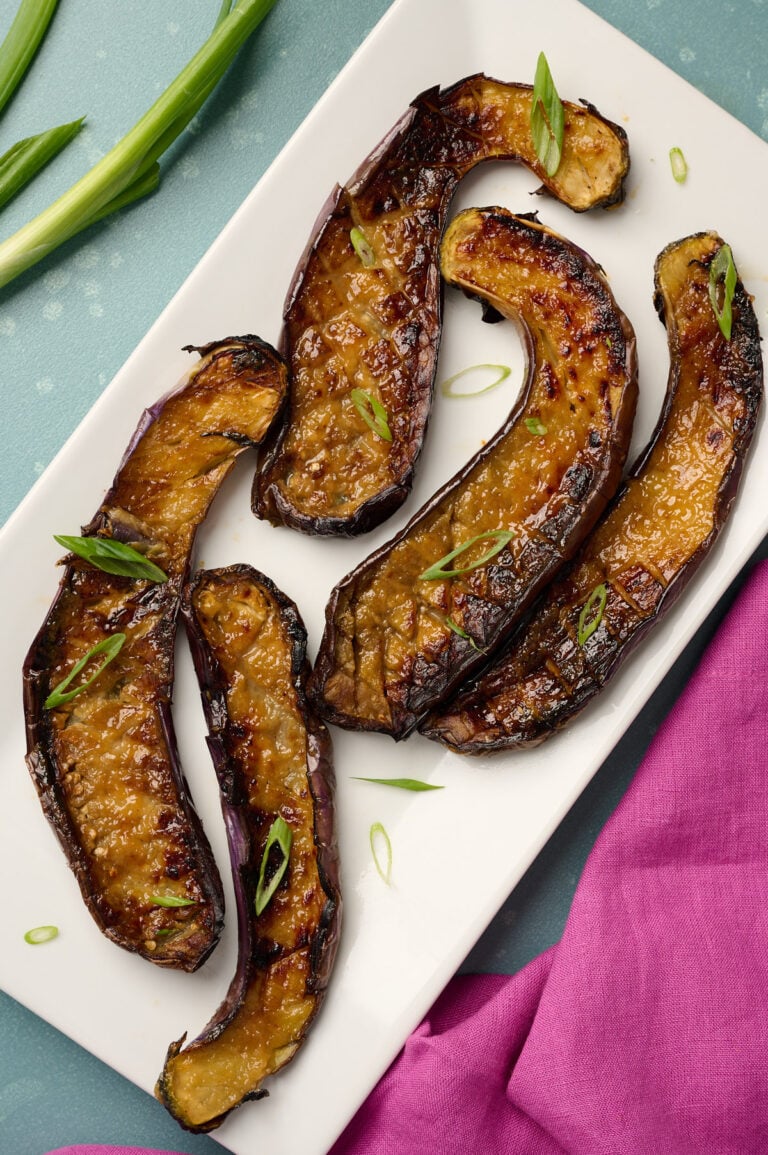 Plated miso eggplant garnished with thinly sliced green onions on a blue surface with pink linen and fresh green onions in the background.