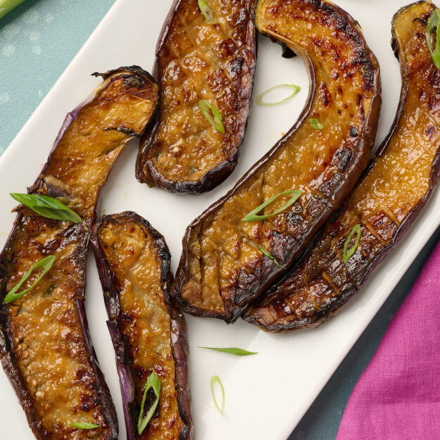 Close-up of plated miso eggplant garnished with green onions, styled on a blue surface with pink linen in the background.