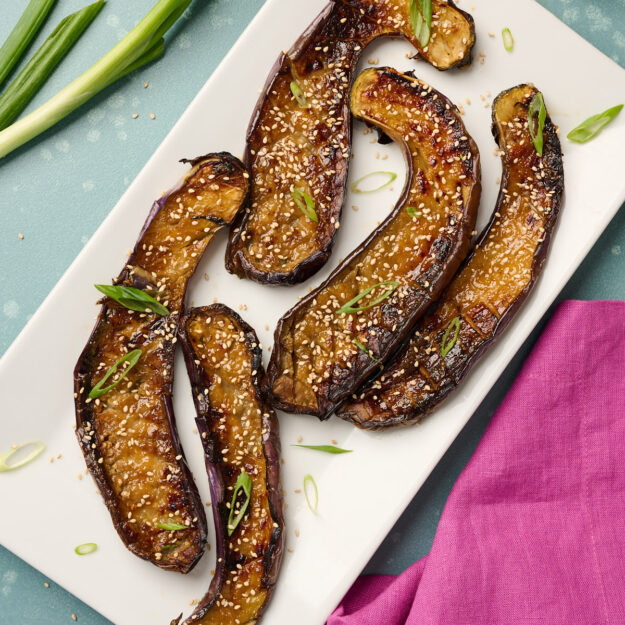 Optional garnish of toasted sesame seeds added to plated miso eggplant with green onions, styled on a blue surface with pink linen and sesame seeds in the background.