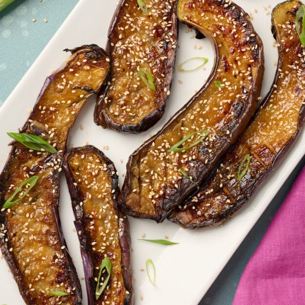 Close-up of plated miso eggplant garnished with toasted sesame seeds and green onions, styled on a blue surface with pink linen.