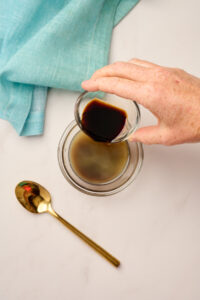 Hand combining soy sauce into yuzu juice in a glass bowl with a gold spoon, styled with blue linen.