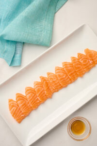 Flat-cut Hira-zukuri salmon slices plated with sesame oil and styled with blue linen.
