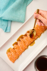 Hand drizzling yuzu soy sauce over salmon sashimi slices with a gold spoon, styled with blue linen.