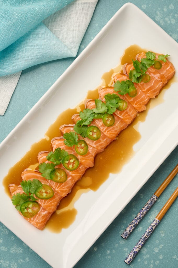 Fully plated salmon sashimi dressed with yuzu soy sauce, garnished with thinly sliced jalapeno and cilantro, styled with blue linen and chopsticks.