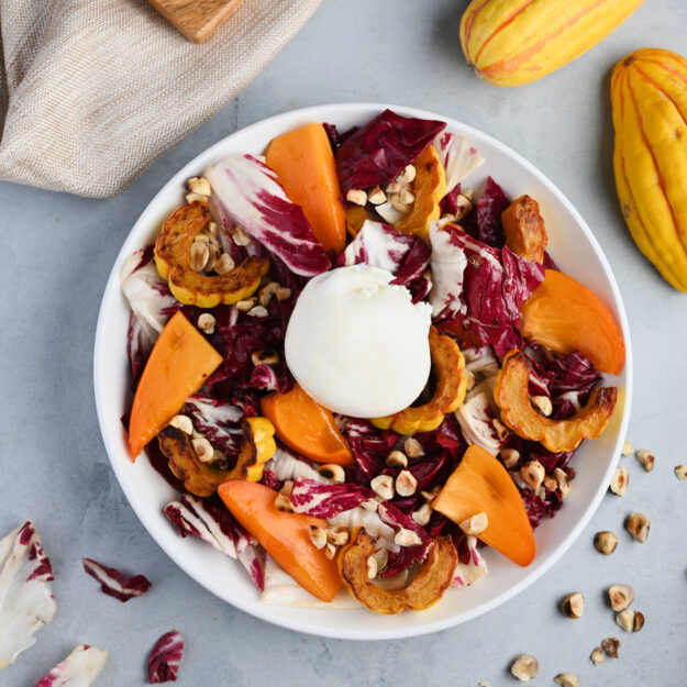 Plated persimmon salad with toasted hazelnuts, roasted Delicata squash, and torn radicchio styled on a beige linen.