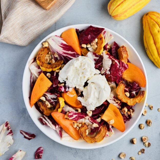 Close-up shot of plated persimmon salad with beige linen, delicata squash, and toasted hazelnuts.