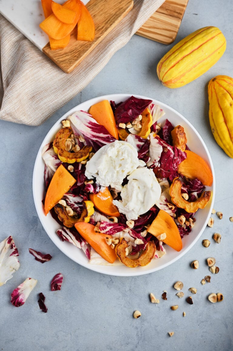 Overhead view of a fully composed persimmon salad with persimmons, roasted delicata squash, hazelnuts, and radicchio, styled with beige linen and wooden cutting boards.