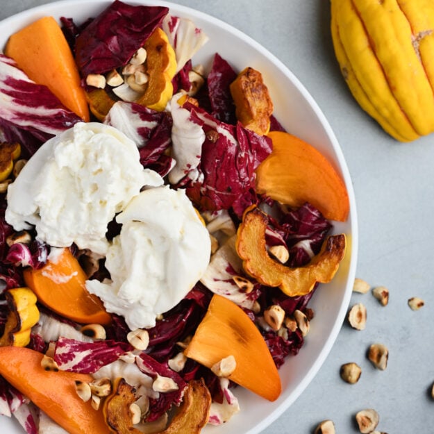 Side shot of the plated persimmon salad with beige linen, Delicata squash, and toasted hazelnuts.