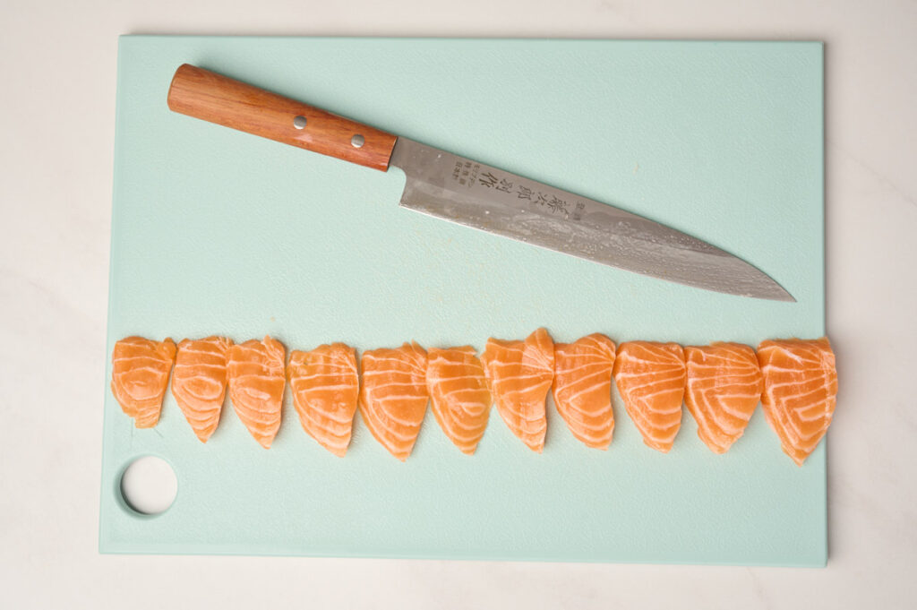 All flat-cut salmon slices, created with the Hira-zukuri method, lined up on a blue cutting board alongside the Yanagiba knife.