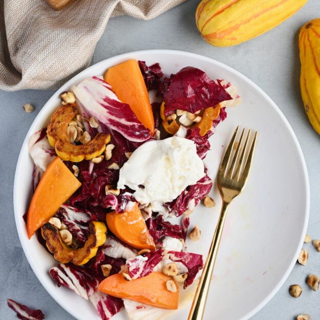 Half-eaten persimmon salad with a gold fork, sliced persimmons, and roasted Delicata squash on a marble cutting board.