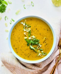 Overhead shot of plated Kabocha squash soup with cilantro, lime, and a gold spoon on a beige linen.