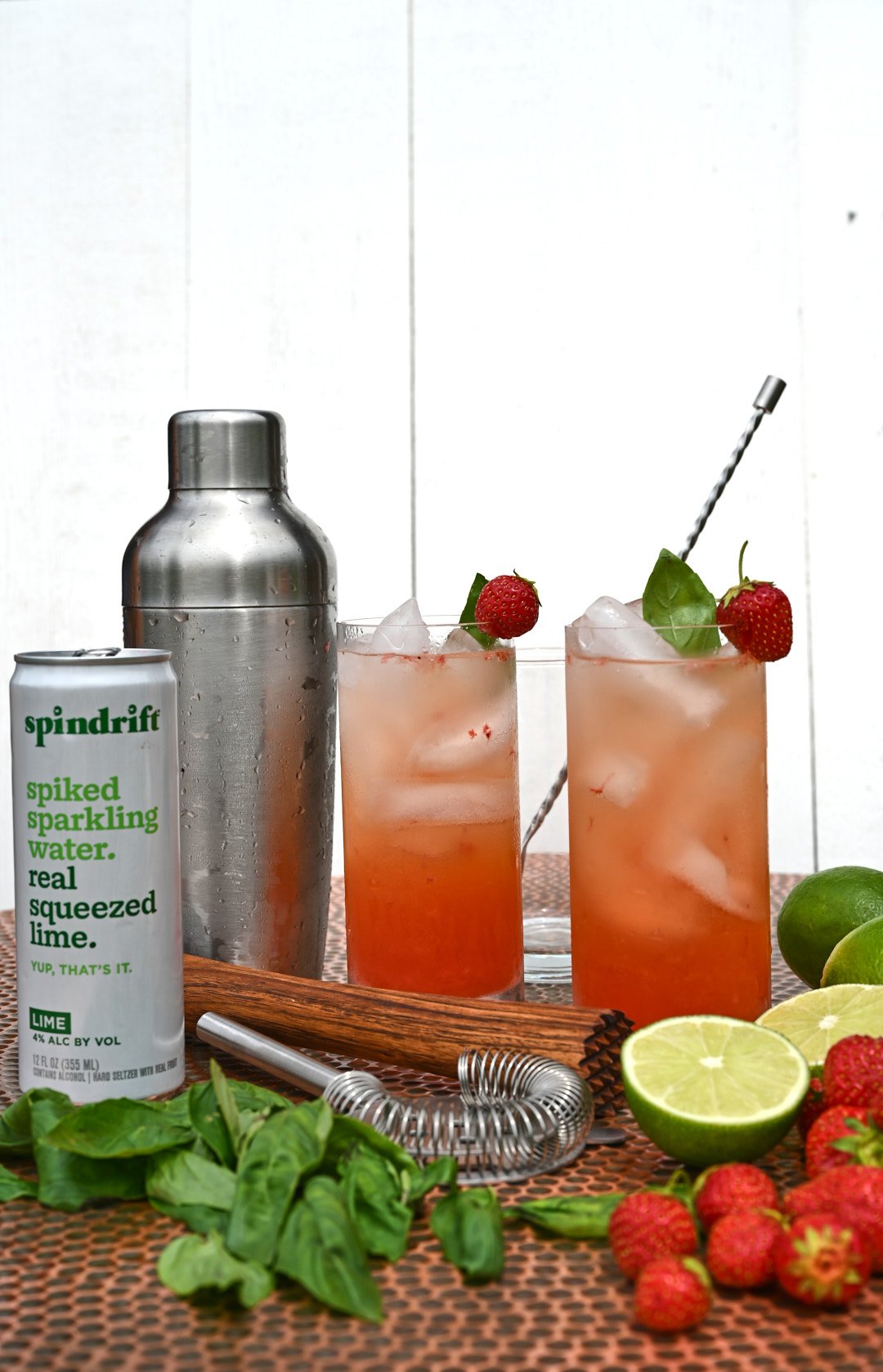 two strawberry cocktails with a shaker, can of sparkling water and limes