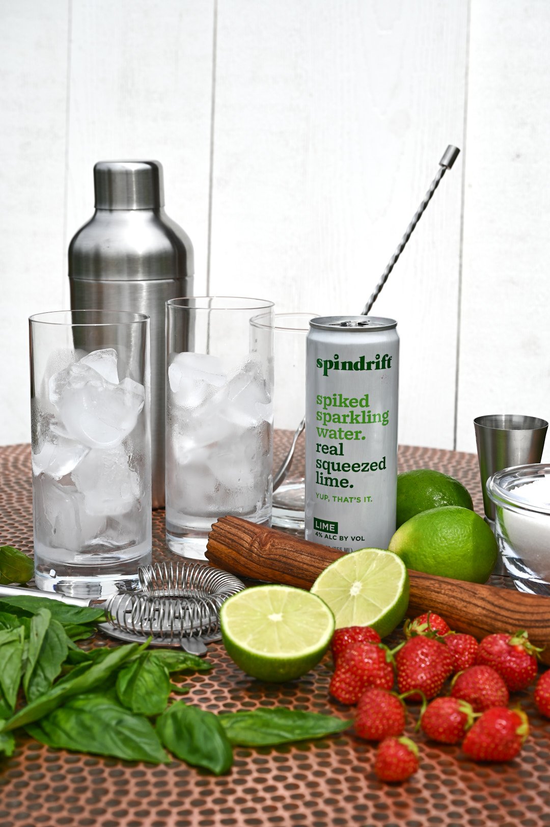 ingredients for cocktail: spindrift sparkling drink, limes, strawberries and mint with iced glasses and cocktail shaker
