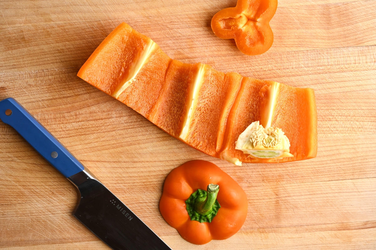 An open pepper on a cutting board in a long strand in the middle of being seeded