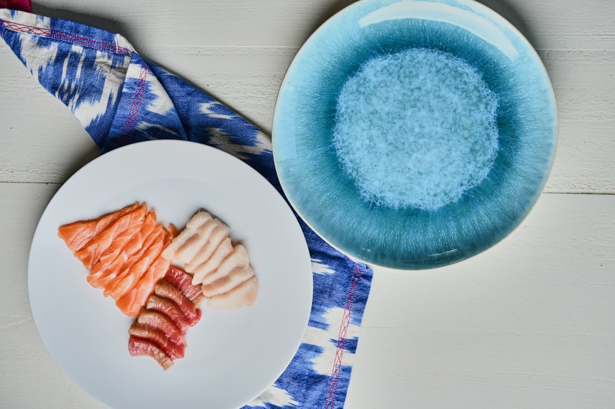 sliced salmon, yellow tail, and toro on a white plate waiting to be plated on a blue decorate plate