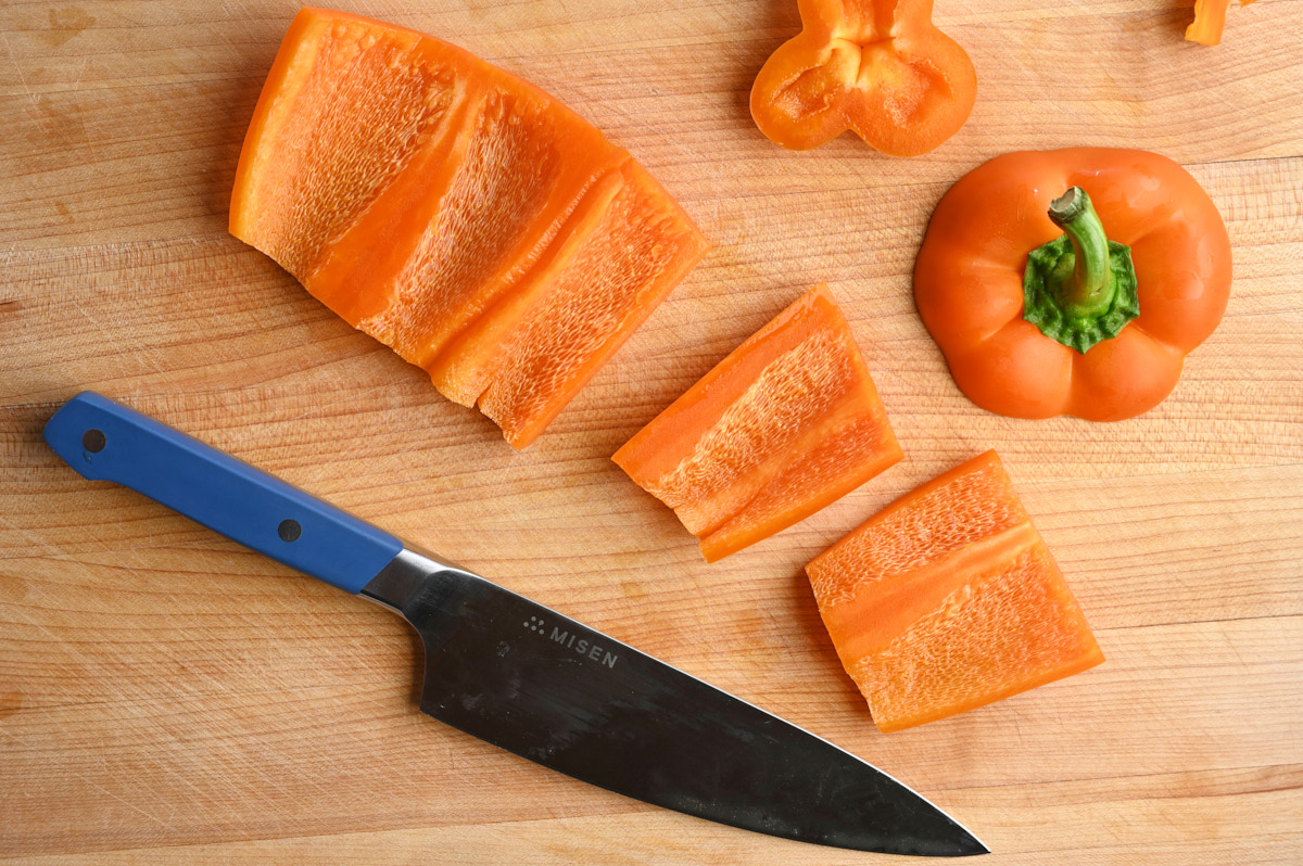 Thick strips of orange bell pepper besides a chef's knife on a wooden cutting board