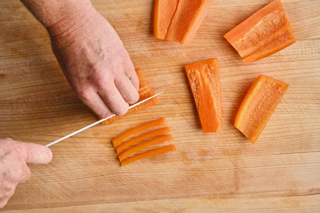 Orange Bell Pepper being cut into small strips on a wooden cutting board