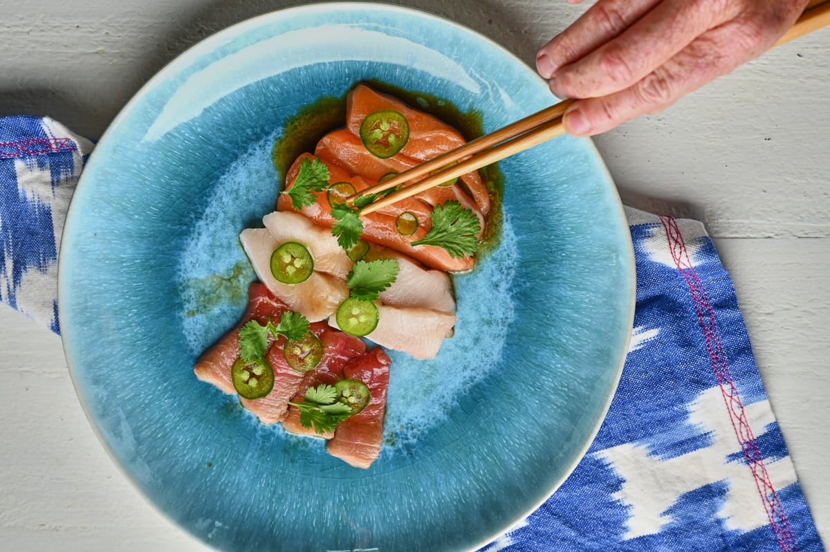 A hand placing Cilantro and jalapenos on Sashimi on a blue plate 