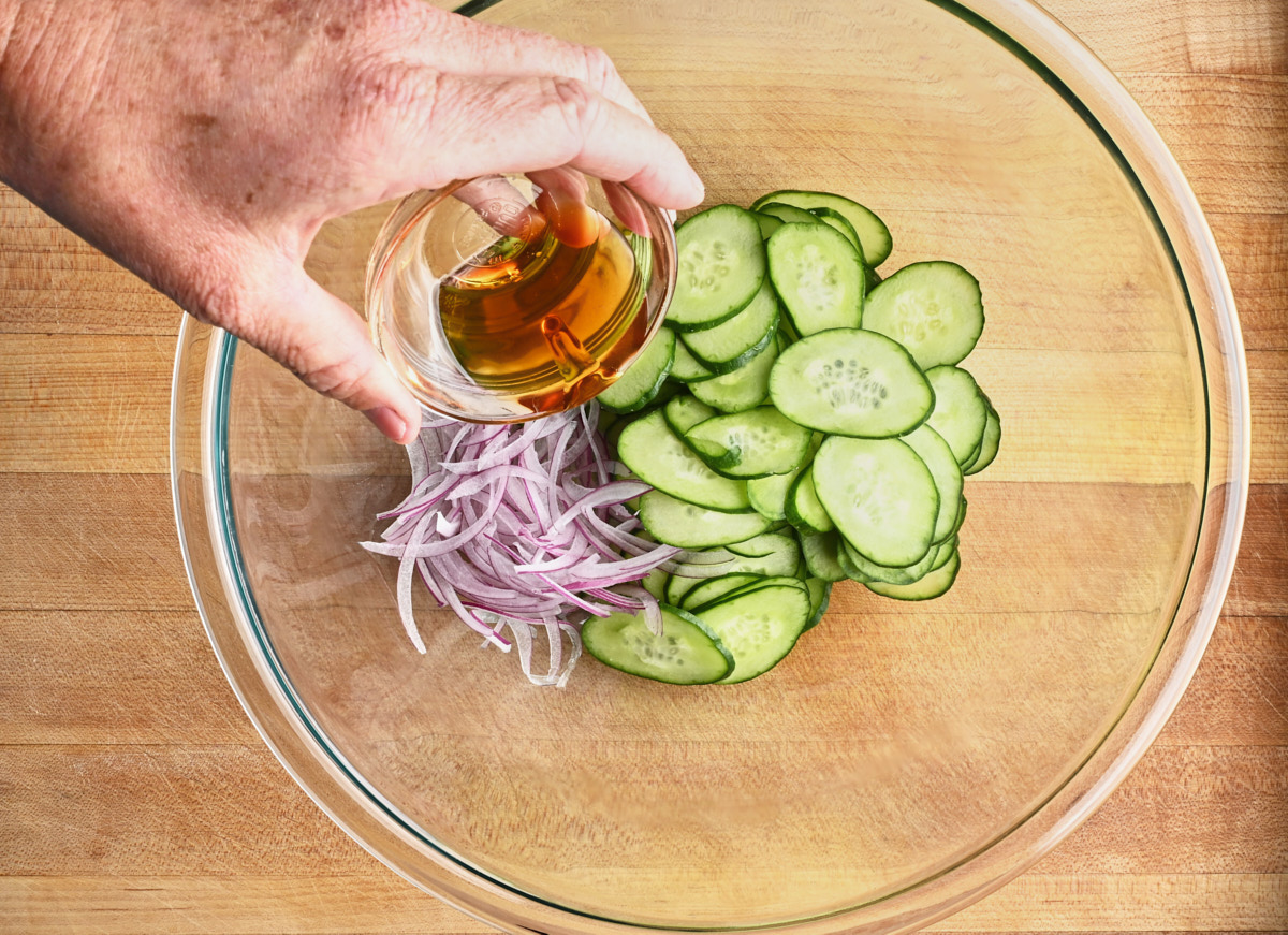 sesame oil being added to cucumbers, and red onion in a glass bowl
