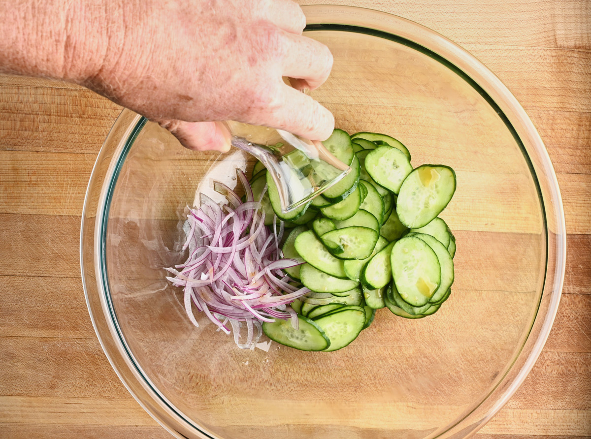vinegar being added to cucumbers, and red onion in a glass bowl