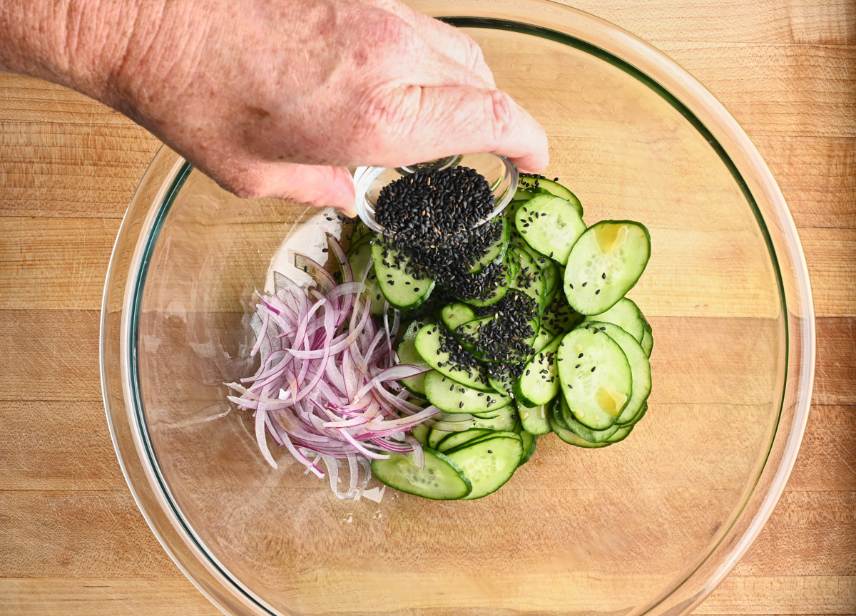 black sesame seeds being added to cucumbers, and red onion in a glass bowl