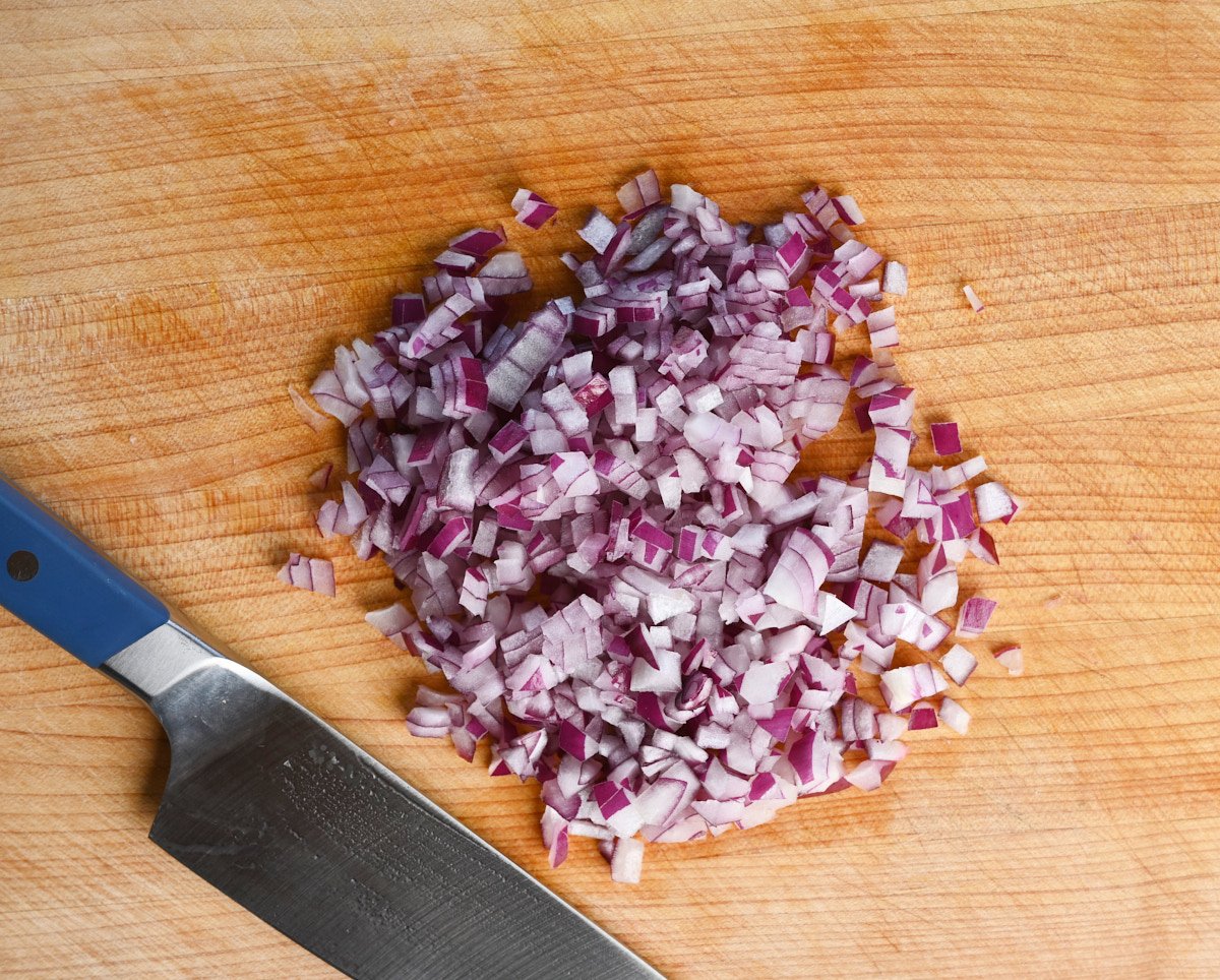 pieced of a diced red onion centered in a pile on a wooden cutting board besides a chef's knife